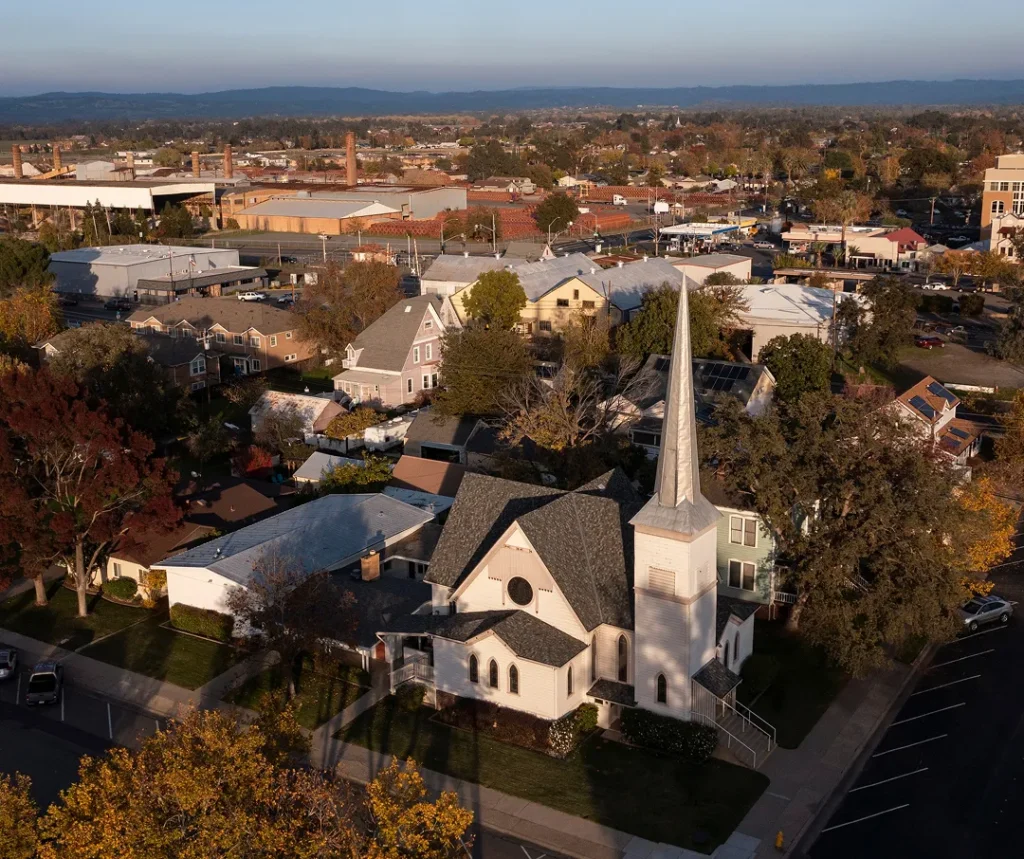 Aerial view of a white church with a tall steeple surrounded by residential and commercial buildings in a suburban area.