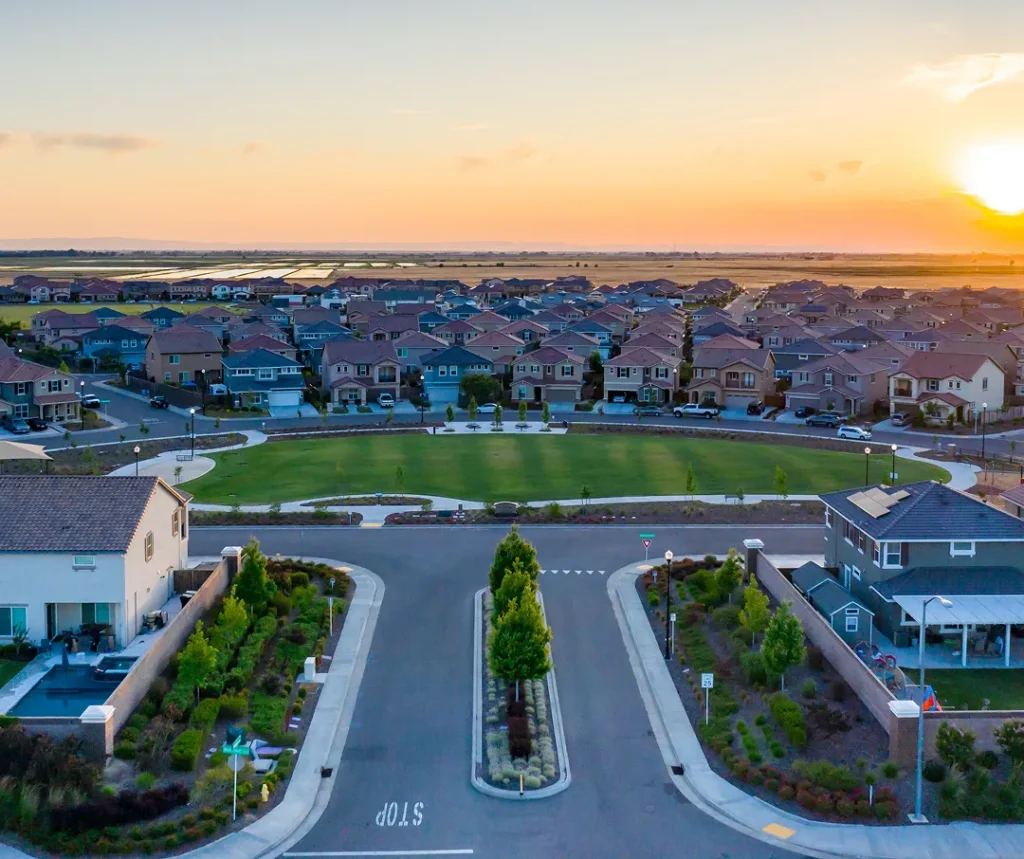 Aerial view of suburban neighborhood with rows of houses, green park, and sunset sky.