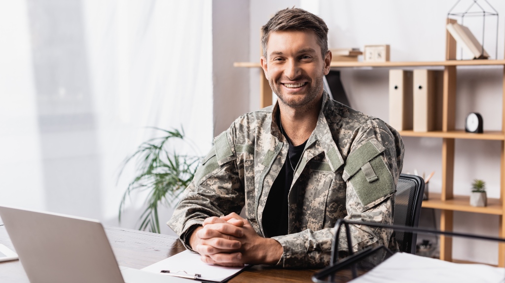 cheerful military man in uniform sitting with clenched hands near laptop on desk